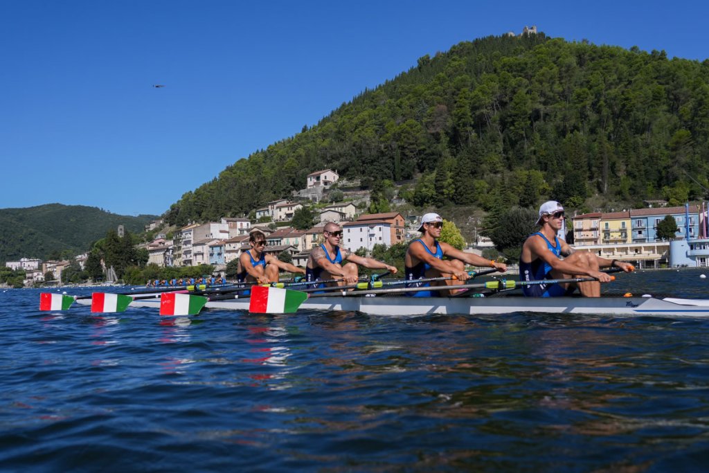 Quattro canottieri italiani in allenamento su un lago, con le bandiere italiane sulle barche e un paesaggio collinare sullo sfondo.