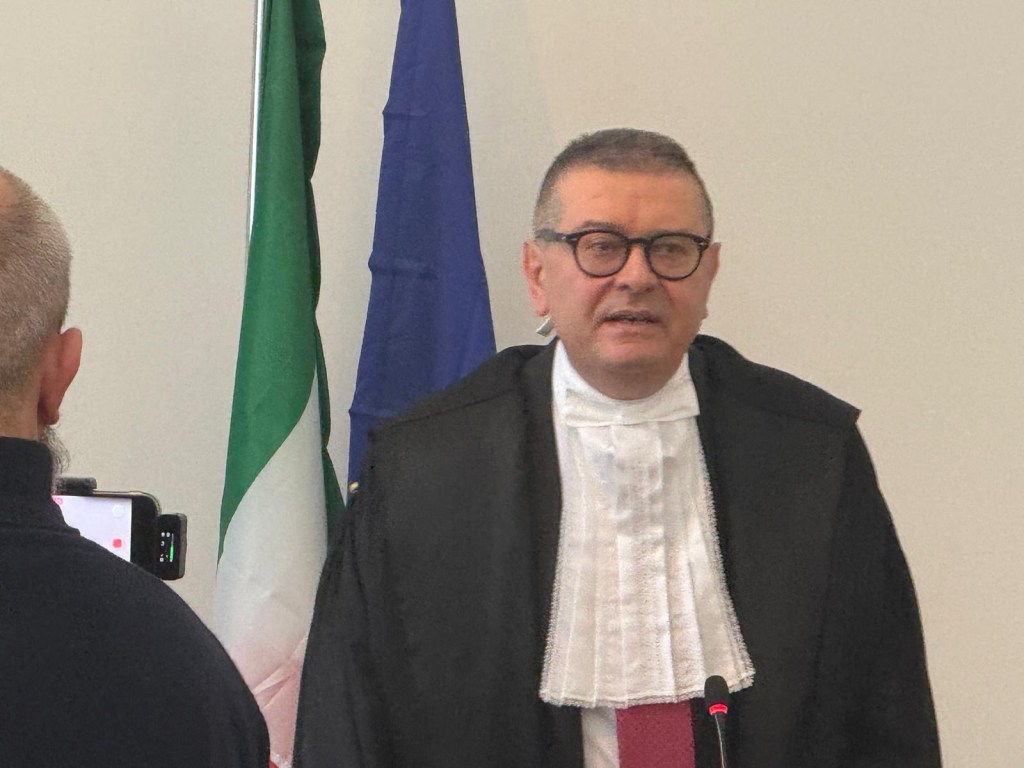 A judge in formal attire speaking at an event, with Italian and European Union flags in the background.