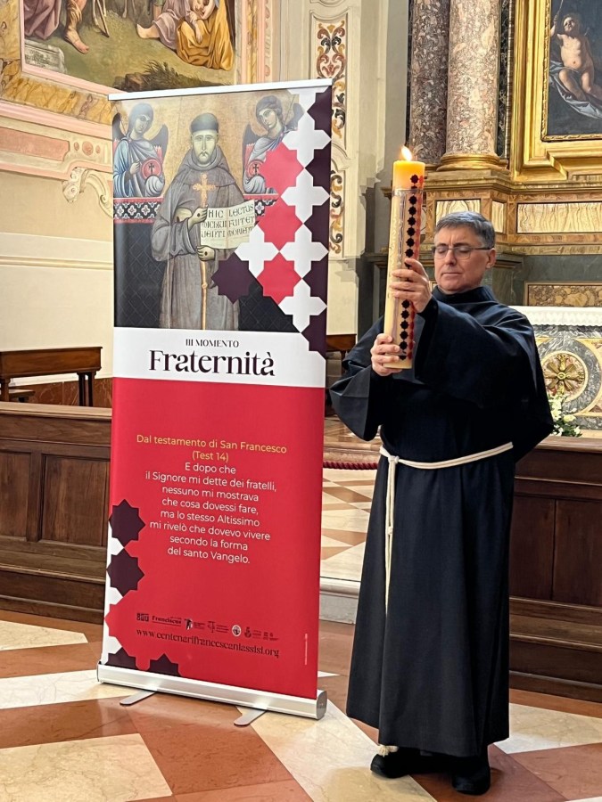 Un religioso in abiti francescani tiene un cero acceso mentre si trova accanto a un banner che celebra il tema della fraternià, all'interno di una chiesa.