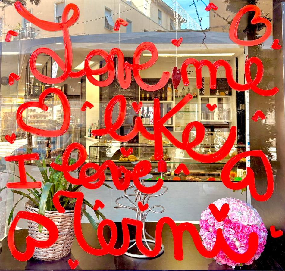 A storefront window with handwritten red lettering that says 'Love me like I love a meri,' surrounded by small heart designs. There is a potted plant and a decorative flower arrangement in the foreground.