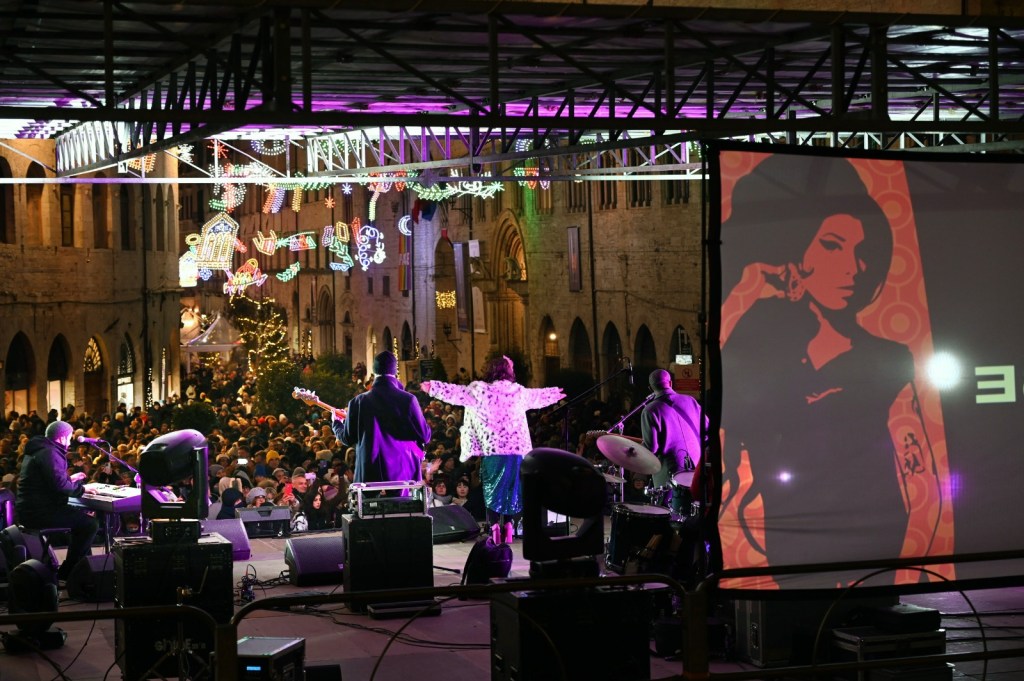 A lively New Year's celebration in Perugia, featuring a live music performance on stage with a large crowd enjoying the festivities below, colorful lights decorating the historic buildings.