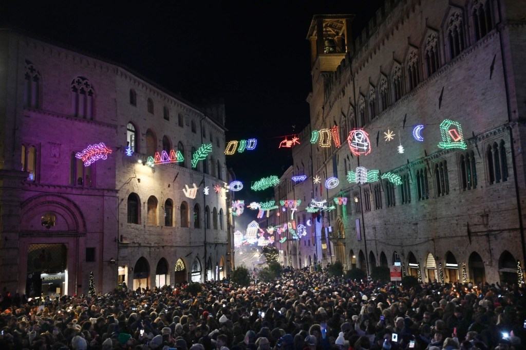 A bustling crowd gathers in the historic center of Perugia during New Year's Eve celebrations, with vibrant holiday lights adorning the buildings and a festive atmosphere.