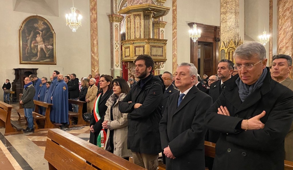 A congregation of people standing in a church, some dressed in dark coats, during a religious service. The interior features ornate decorations and a large painting on the wall.