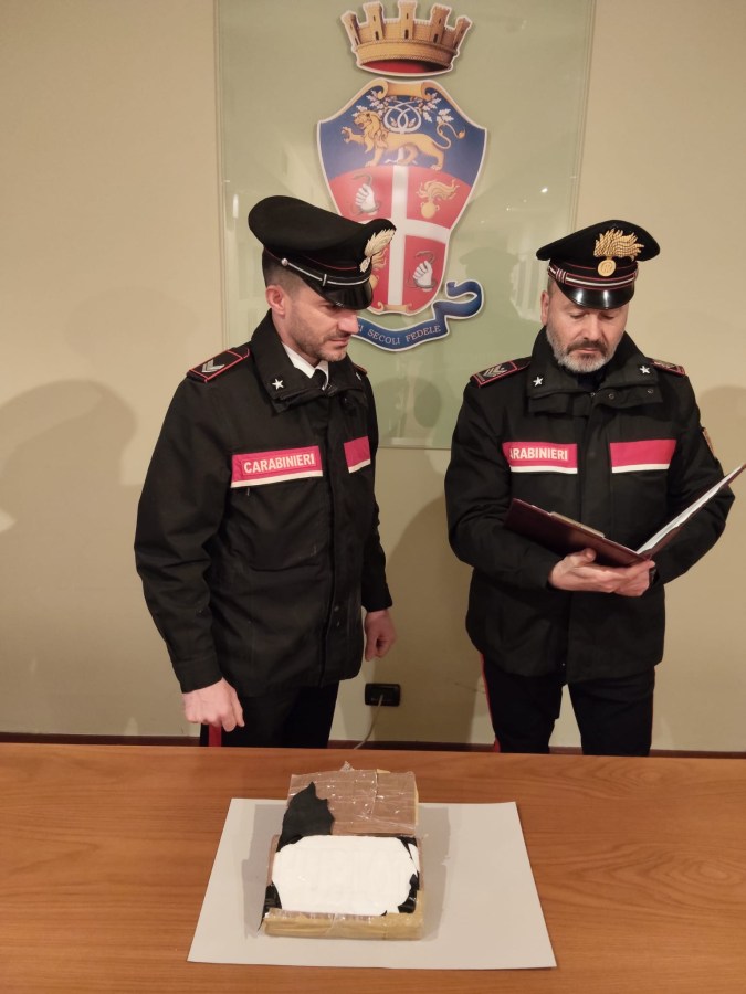 Two Carabinieri officers examining a rectangular package on a table, with one officer reading from a folder while the other observes. A coat of arms is displayed in the background.