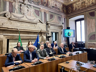 A group of people seated at a conference table in an ornate room, with flags and a presentation screen visible in the background.