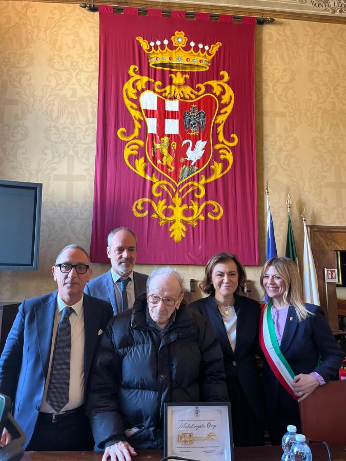 A group of five people stand in front of a large red banner featuring a coat of arms. One elderly man, dressed in a black coat, is seated in front of a certificate commemorating him. The others, including three men and one woman, are dressed in formal attire. The setting appears to be an official or ceremonial space.