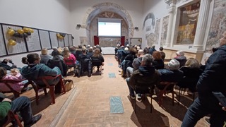 Audience seated in a hall watching a presentation on a screen, with decorative walls and a large window in the background.