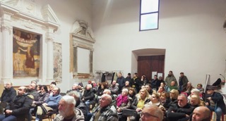 A large audience seated in a hall with historical decor, attentively listening to a presentation.