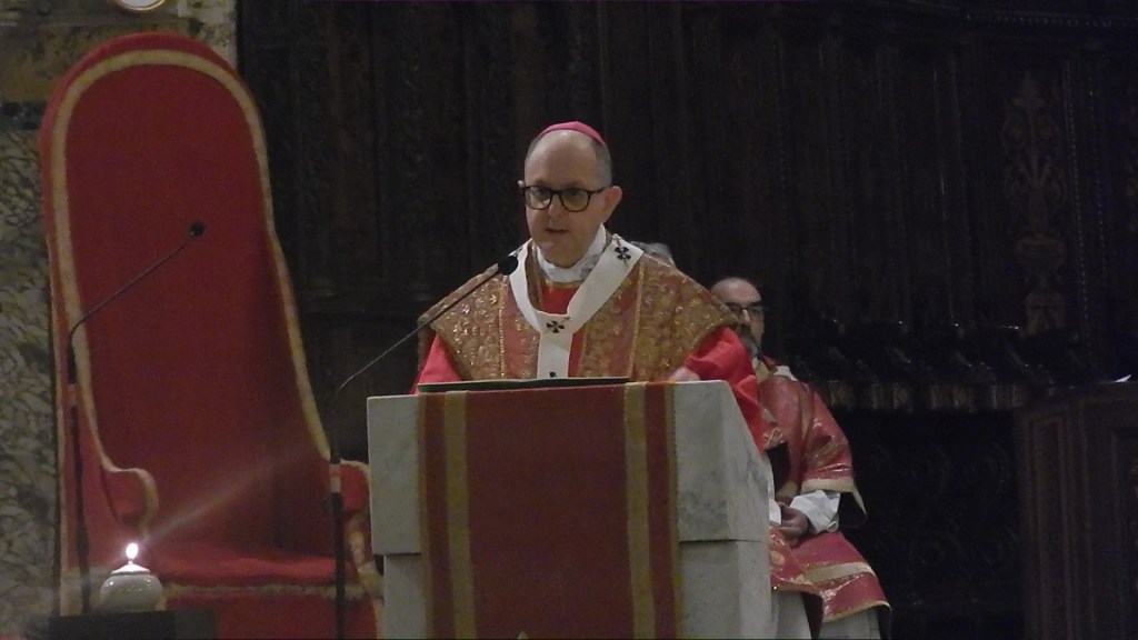 A bishop in ceremonial vestments delivering a sermon at a church altar, with ornate decorations in the background.