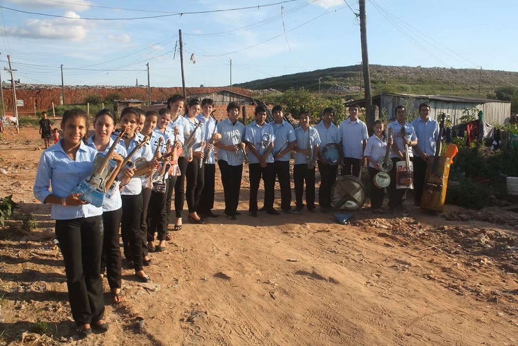 A group of musicians dressed in light blue shirts standing outdoors, holding various musical instruments in a rural setting.