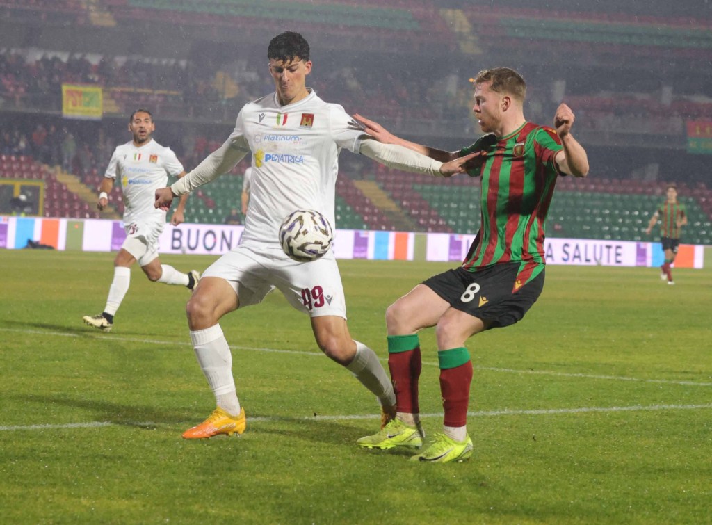 Giocatori di calcio in azione durante una partita, con uno che tenta di affrontare l'avversario in un campo bagnato dalla pioggia.