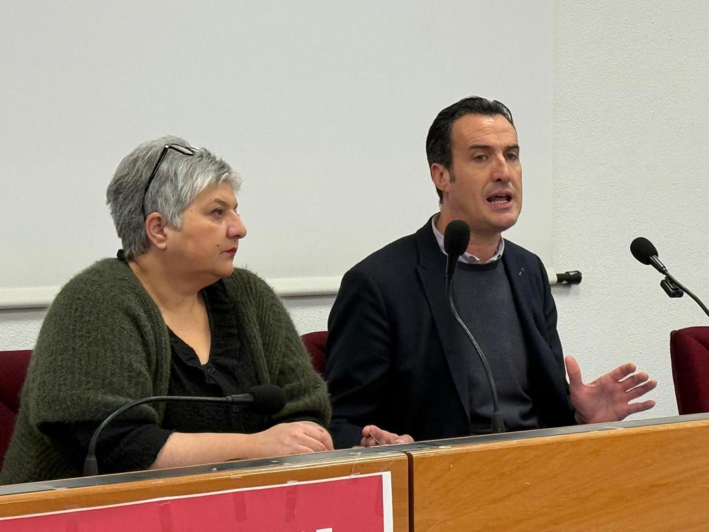 Due relatori durante una conferenza stampa, uno dei relatori parla mentre l'altra ascolta, con un tavolo in legno e un pannello rosso visibile sullo sfondo.