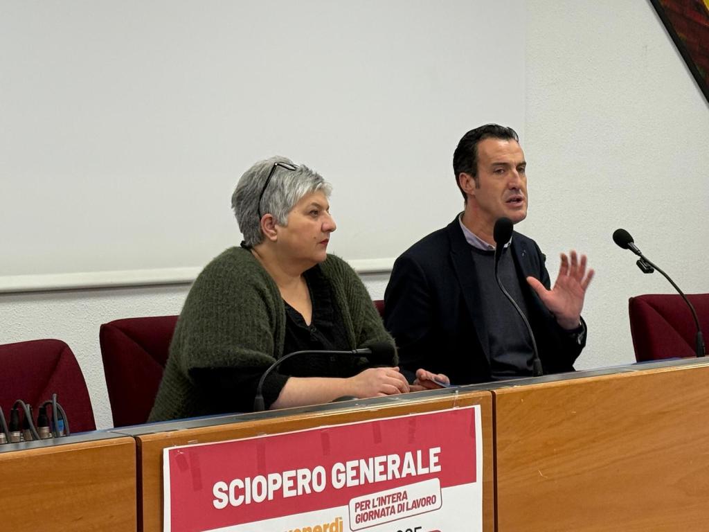 Due rappresentanti della Cgil durante una conferenza stampa, seduti a un tavolo con un cartello che annuncia uno sciopero generale sullo sfondo.