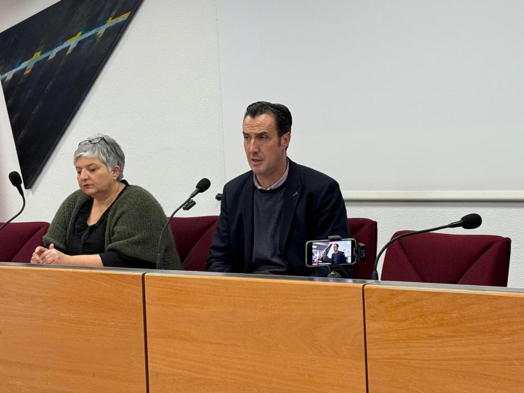 Due persone sedute di fronte a un tavolo durante una conferenza stampa, con un microfono di fronte a ciascuna. Una persona indossa un maglione verde e ha i capelli grigi, mentre l'altra indossa un abito scuro e parla. Un telefono cellulare è posizionato sul tavolo di fronte a loro.