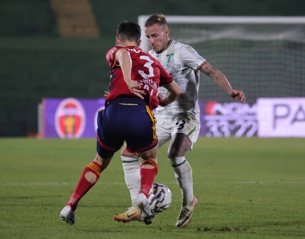 Two soccer players compete for the ball on the field, one in a red and yellow jersey and the other in a white jersey.
