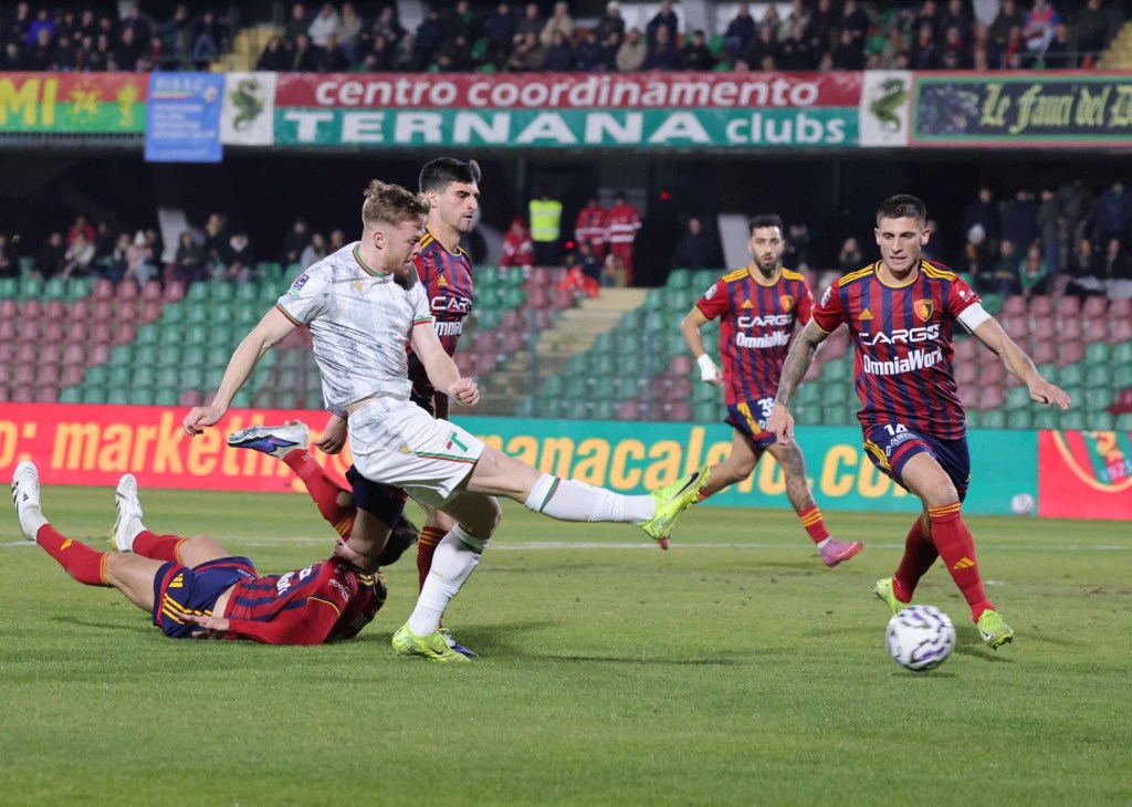 A soccer match in progress with players from two teams competing for the ball. One player is kicking the ball while another is on the ground. Spectators can be seen in the background.