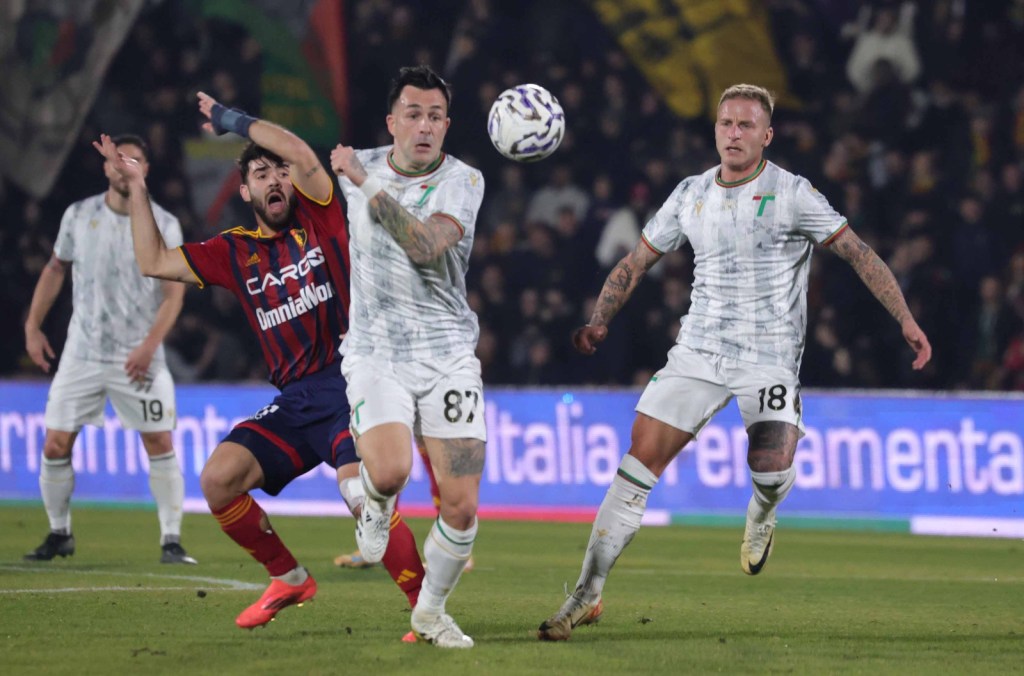 A football match scene with players in action on the field. One player in a red and yellow jersey is attempting to challenge another player in a white and green jersey who is poised to kick the ball. Two other players in the background are observing the play.