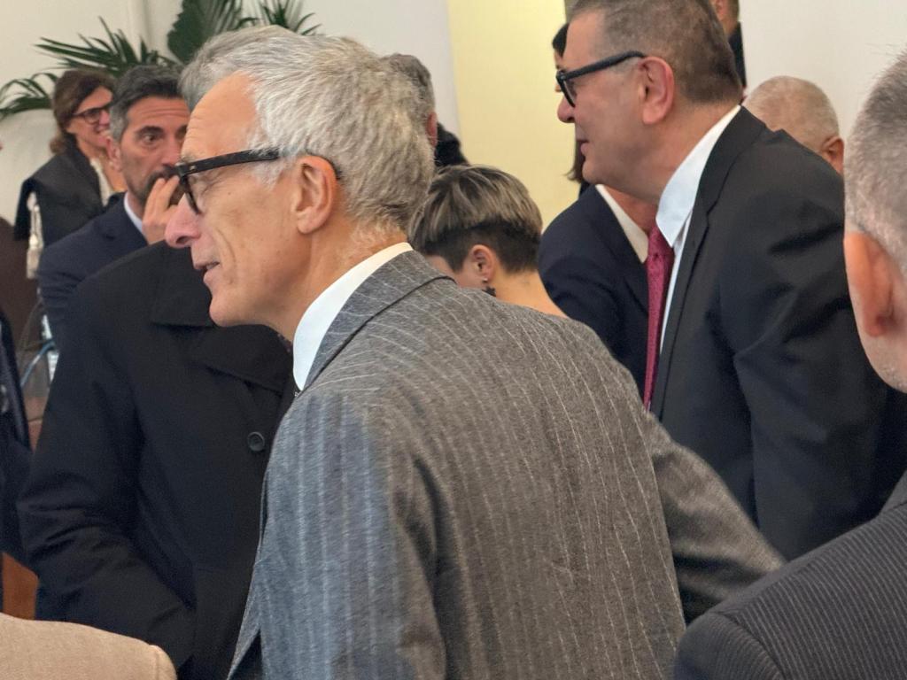 A group of people in a business setting, with one man in a gray striped suit speaking, surrounded by others in formal attire.