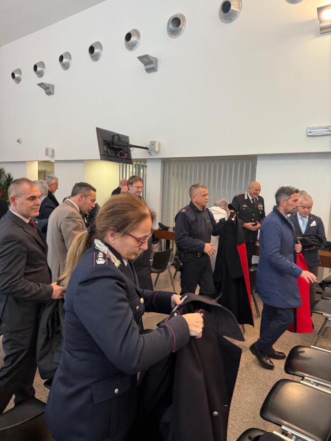 A group of people in formal attire gathered in a bright interior space, interacting while putting on or adjusting coats, with some individuals in police uniforms.