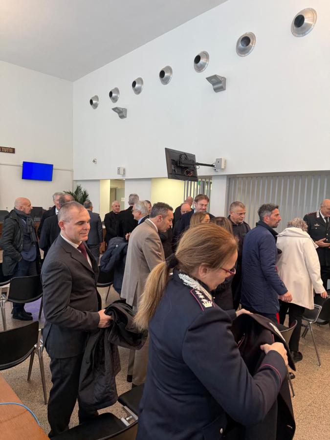 A group of people, some in formal attire, are gathered indoors, mingling and standing near chairs in a well-lit room with modern decor. There is a large screen on the wall and several people are seen holding jackets.