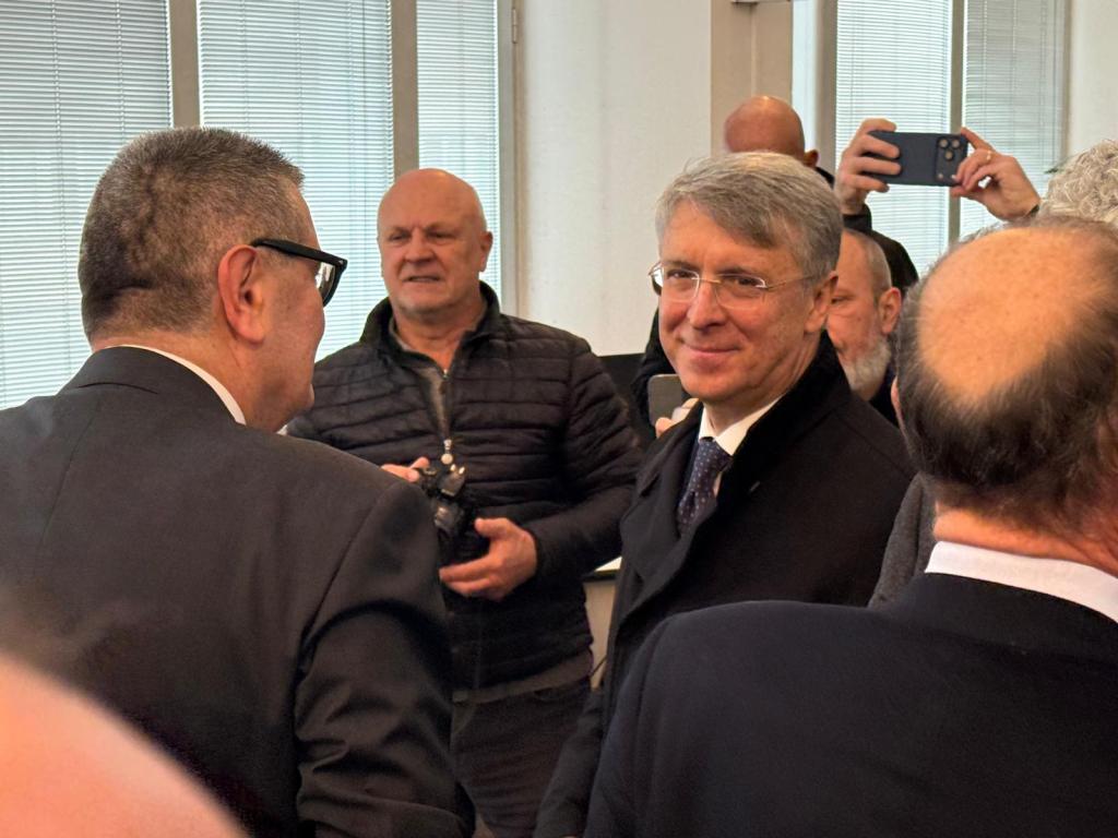A group of people engaged in conversation indoors, with one man in a suit smiling and looking at another man wearing glasses. Several other attendees are visible in the background, some taking photographs.