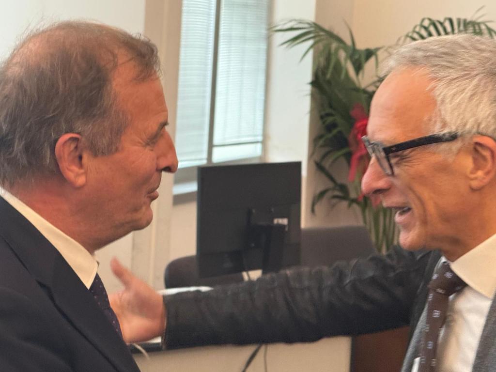 Two men engaging in a friendly conversation indoors, smiling and gesturing. A plant and computer are visible in the background.