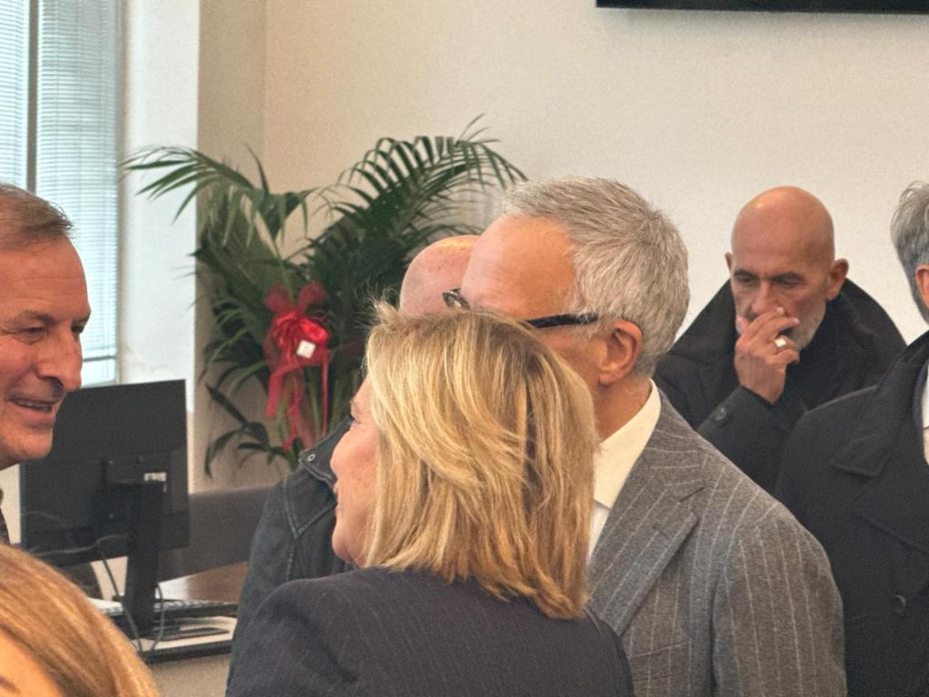 A group of people engaged in conversation indoors, with a palm plant in the background and a decorative element visible. Two individuals are prominently talking, showing friendly interaction.