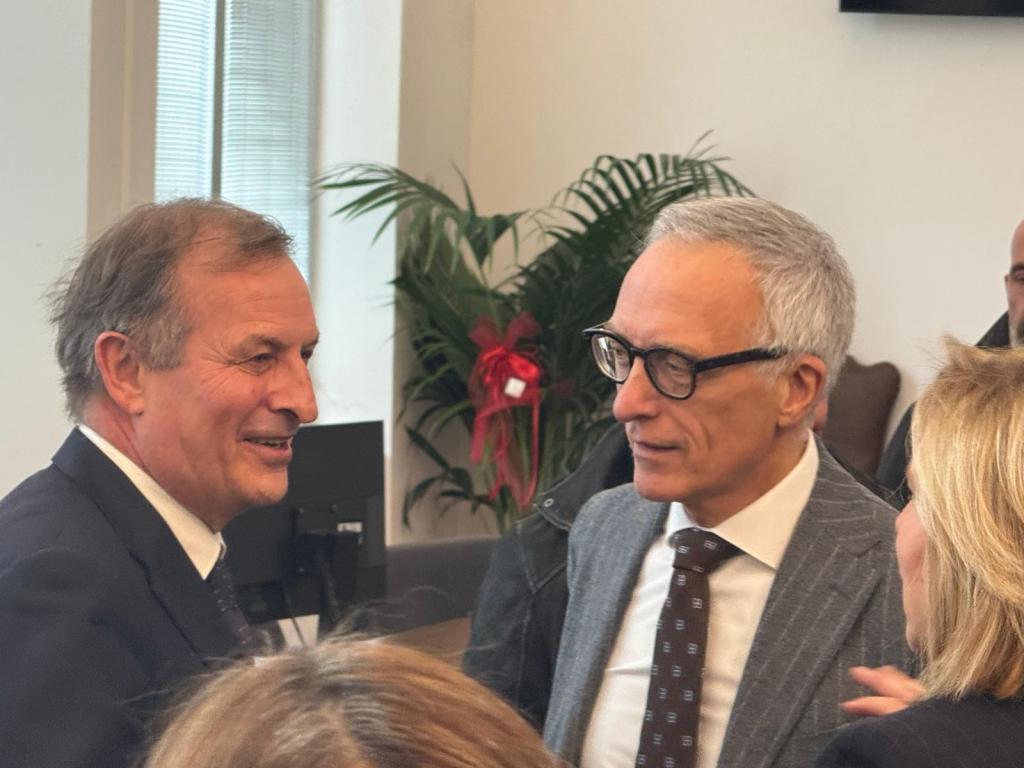Two men smiling and engaging in conversation in a well-lit indoor setting with greenery in the background.