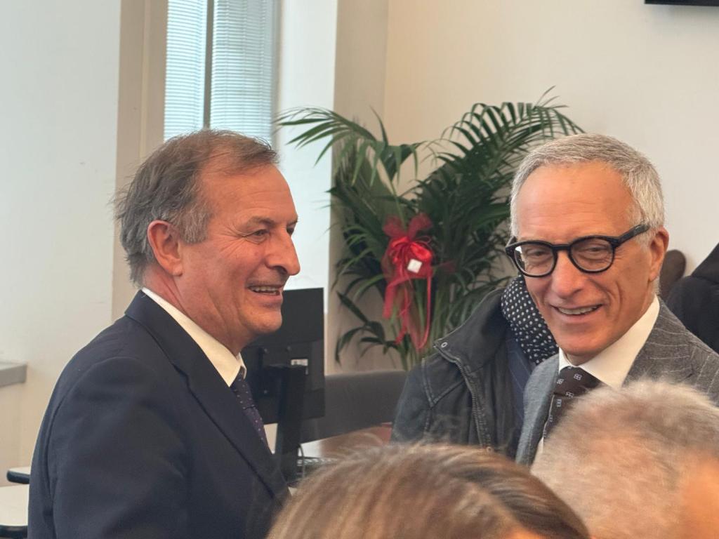 Two smiling men engaging in conversation in an indoor setting, with a plant in the background.