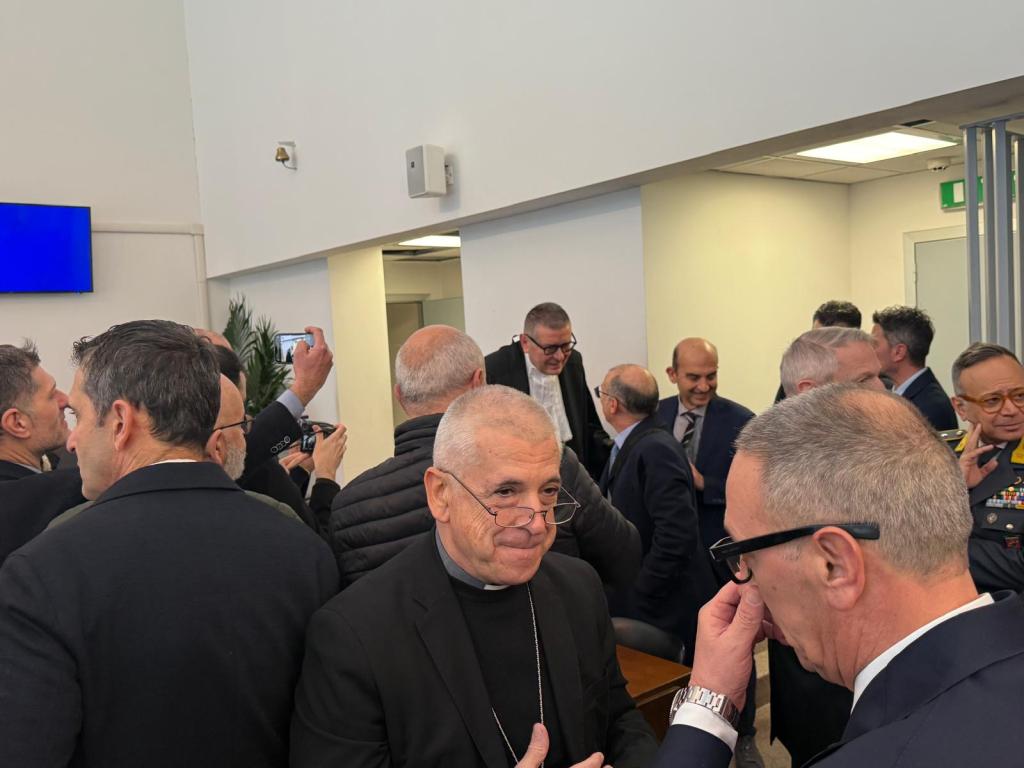 A crowded indoor setting where several men in formal attire are engaged in conversation. Some individuals appear to be greeting each other while others are using their phones.