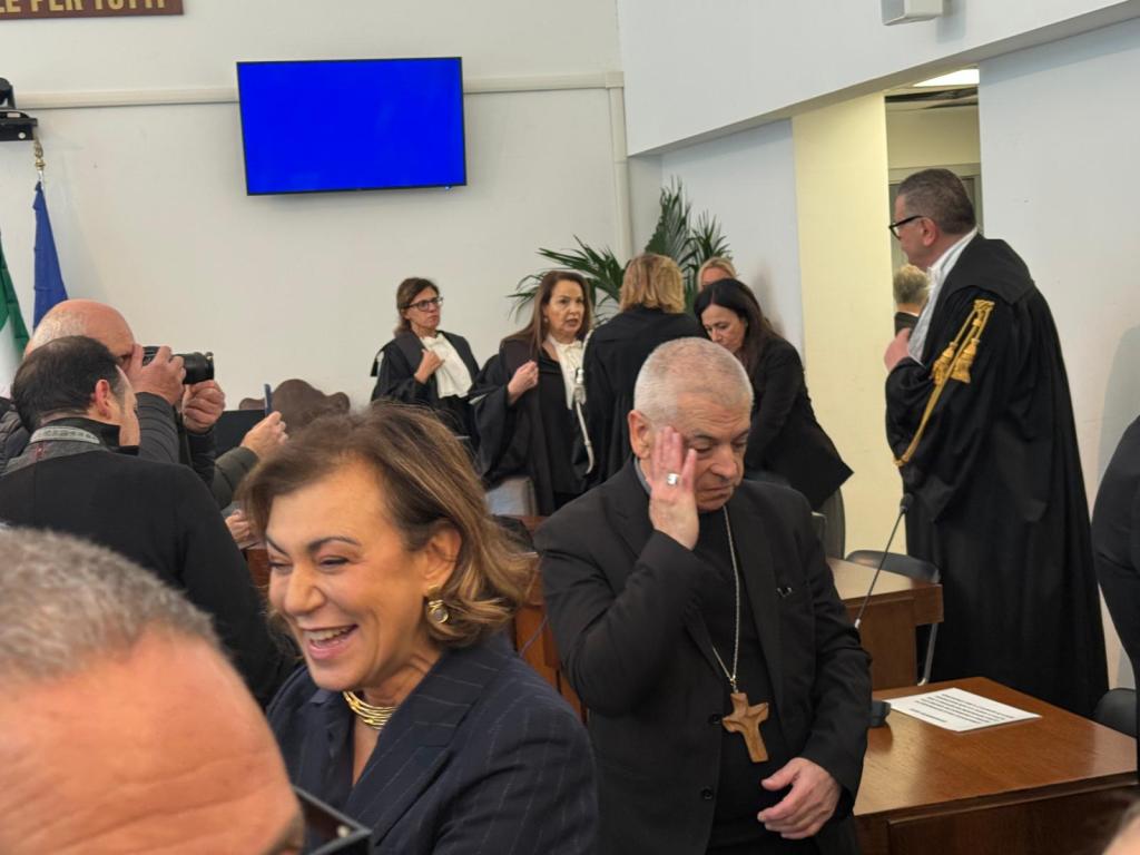 A group of people, including a man wearing a cross necklace, gather in a courtroom setting, with a few individuals in formal attire discussing and others taking photographs.