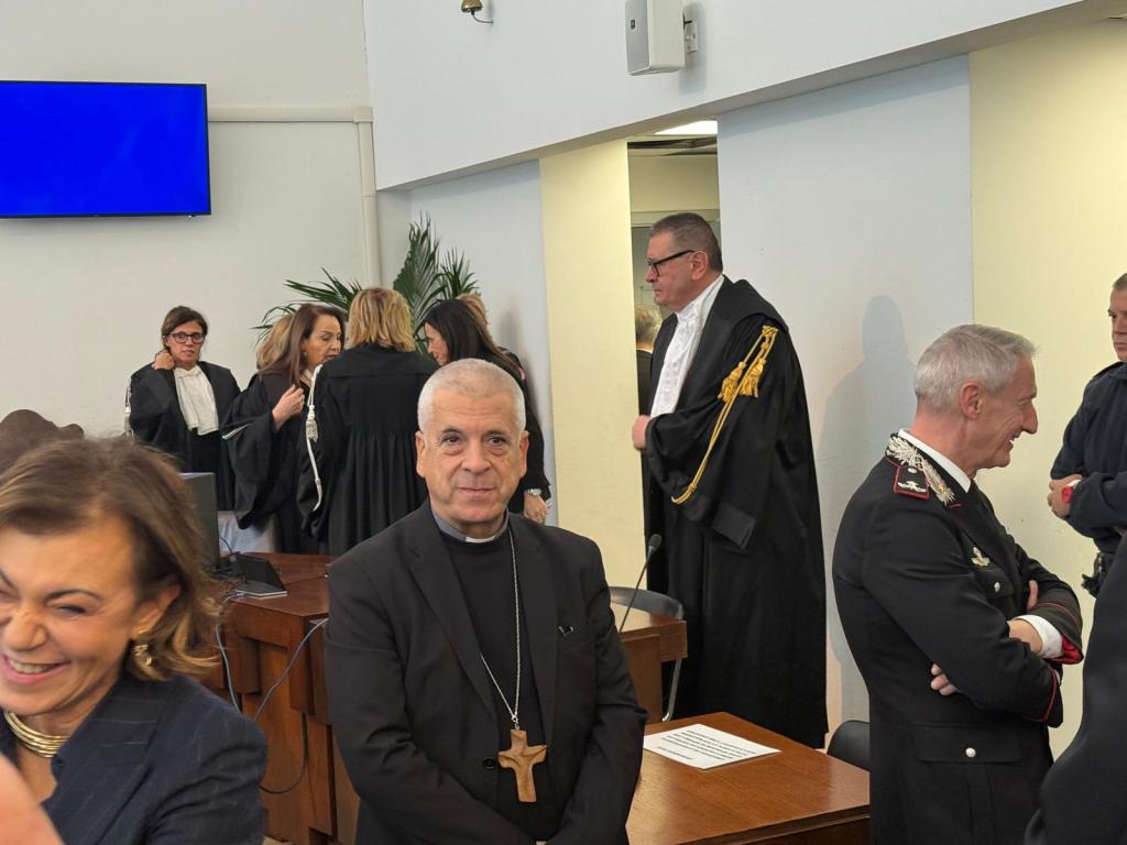 A group of individuals dressed in formal attire, including judges and legal professionals, gathered in a courtroom setting. In the foreground, a man wearing a clerical collar and a cross stands next to a laughing woman, while others are engaged in conversation in the background.