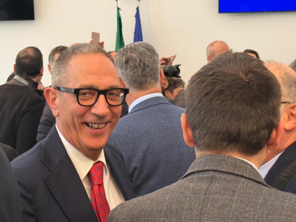 A smiling man with glasses, dressed in a suit, stands among a crowd of people at an event. He appears engaged and friendly.