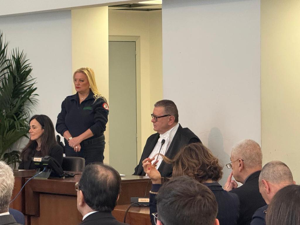 A courtroom scene featuring a judge in black robes, an officer standing, and several people seated in the background. The setting includes a wooden bench and green indoor plants.