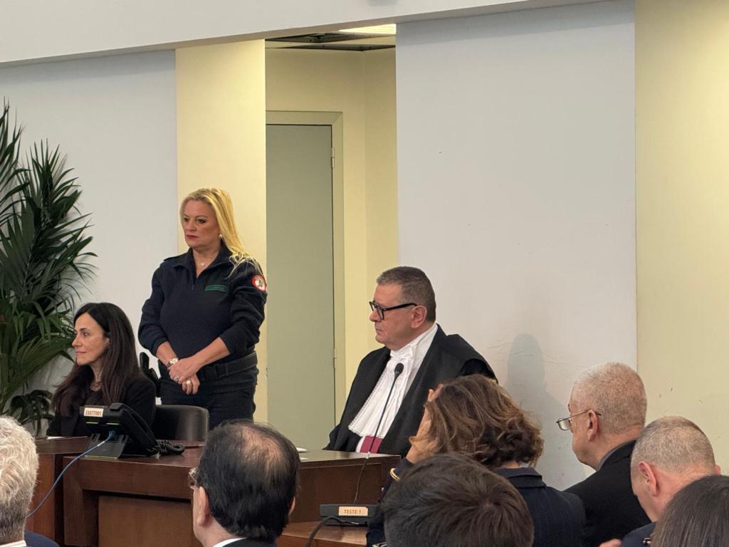 A courtroom scene featuring a female officer standing next to a table with legal professionals, including a judge in formal attire, while audience members are visible in the foreground.