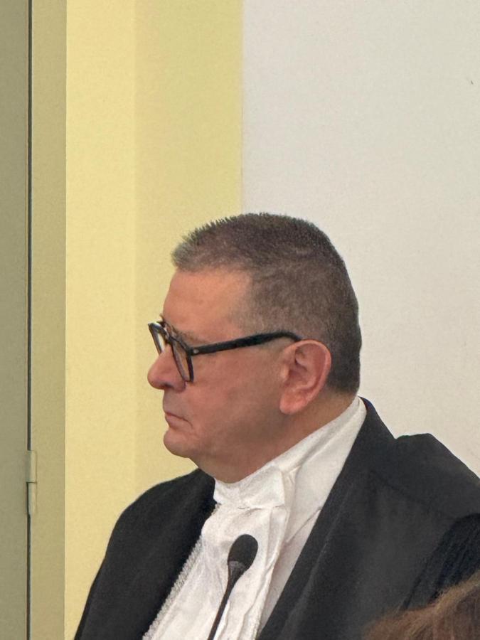 A middle-aged man with glasses wearing a black robe and white collar, appearing thoughtful while standing next to a microphone.