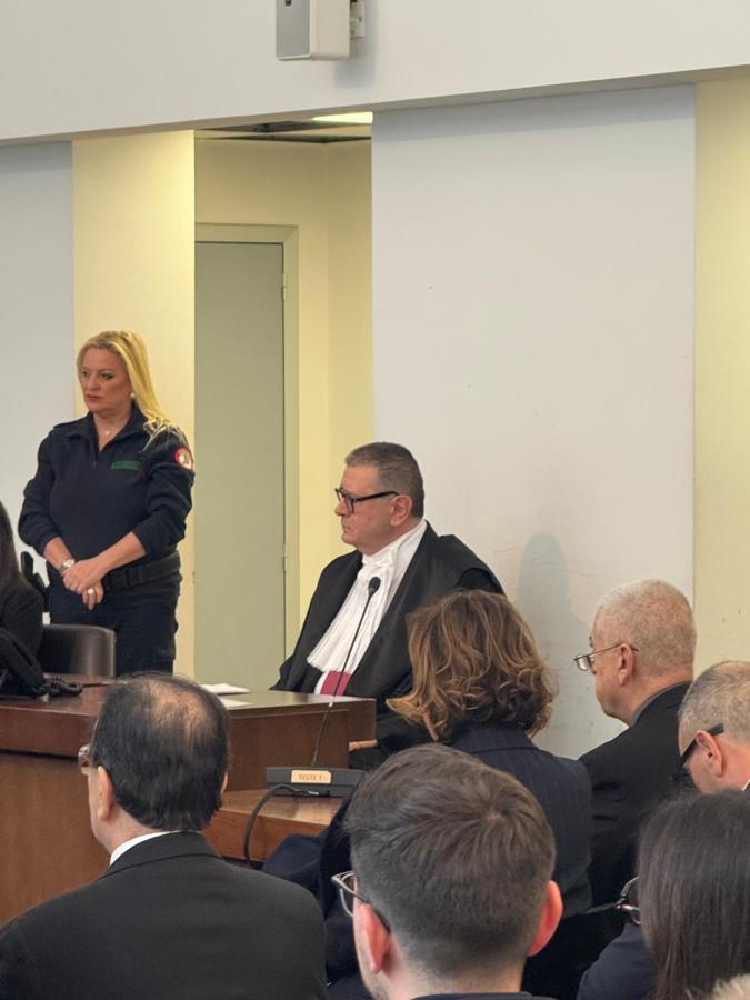 A courtroom scene with a judge in formal attire seated at a bench, a female officer standing nearby, and various attendees in the foreground, including lawyers and other individuals.