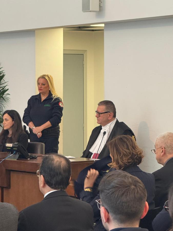 A courtroom scene featuring a woman standing in a uniform, a judge in robes, and several attendees seated in the foreground.