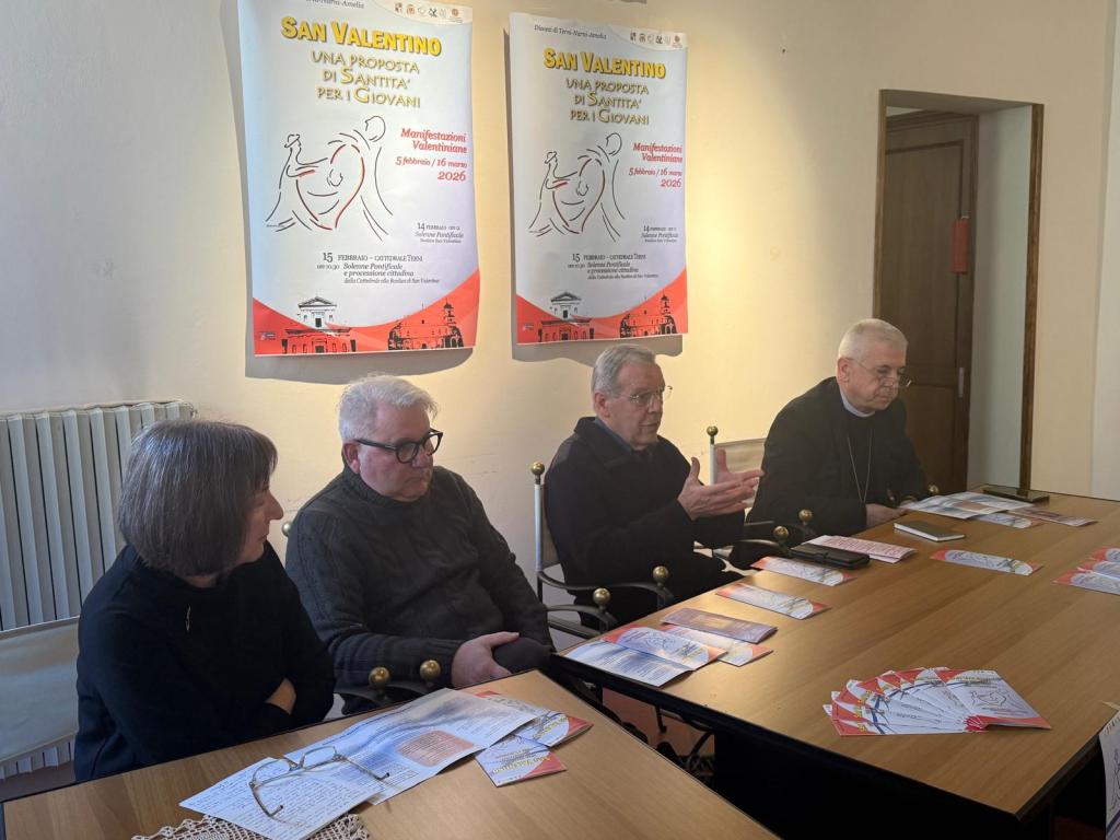 A group of four individuals seated at a table discussing a community event. Behind them are two posters related to a 'San Valentino' event scheduled for February 2026, promoting a message of holiness for youth.