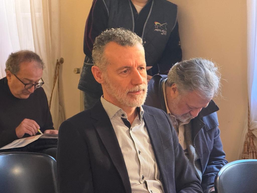 A man with salt-and-pepper hair and a beard, wearing a blazer and checked shirt, sits in a seating area. He appears focused, while other men in the background are engaged in writing or looking down.