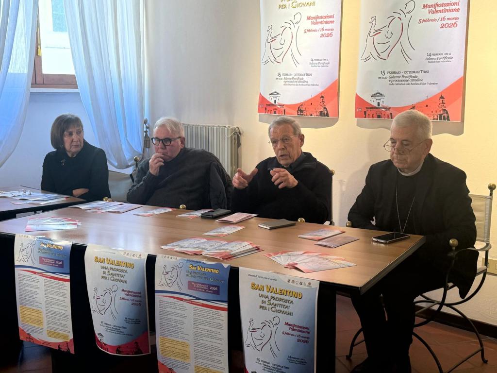A group of four individuals seated at a table with promotional materials about an event related to Valentine celebrations in 2026. Two are engaging in conversation, with posters displayed in the background.