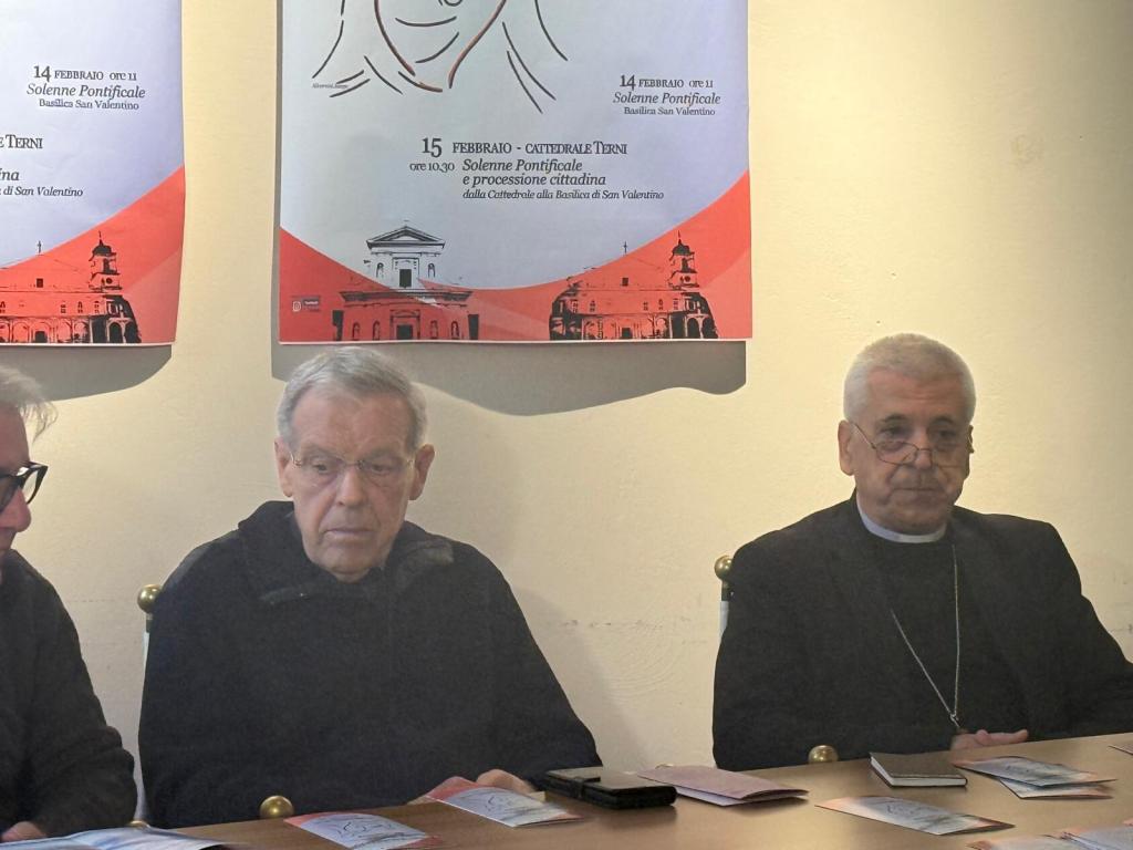 Two men sitting at a table during a press conference, with a poster on the wall announcing events related to San Valentino.