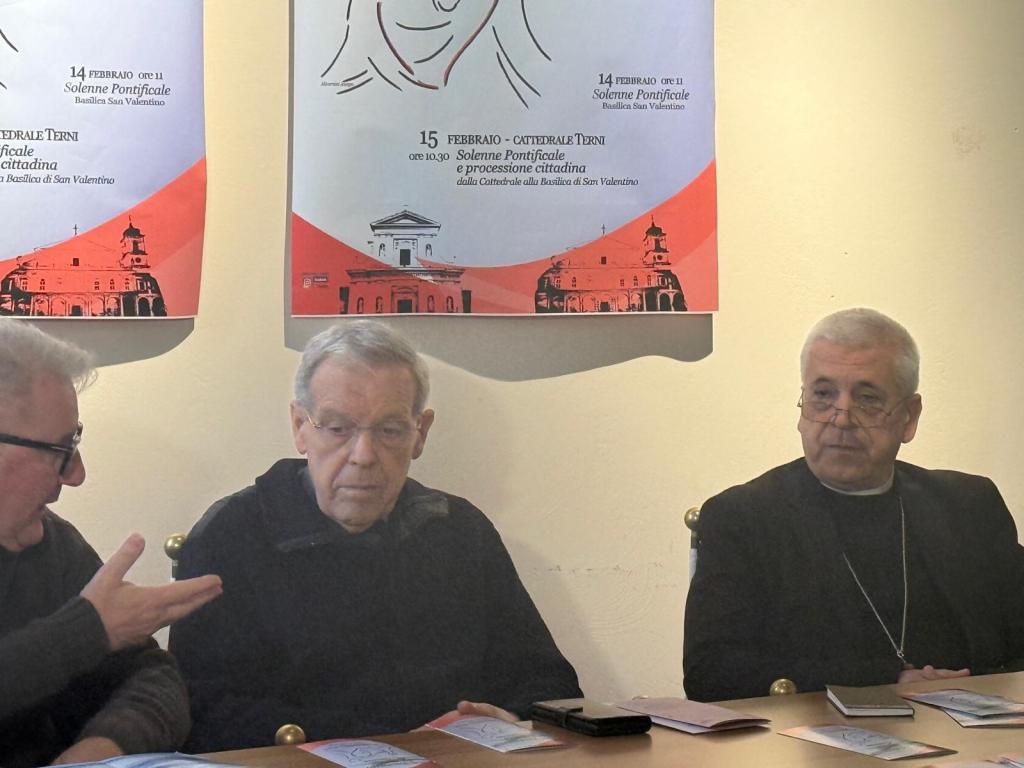 Two elderly men seated at a table, engaged in conversation; one is gesturing with his hand. Poster outlines upcoming religious events in the background.