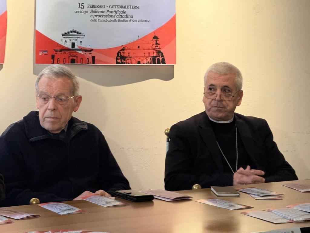 Two men seated at a table with pamphlets in front of them. One man has gray hair and glasses and is dressed in a black sweater, while the other man has white hair, glasses, and is wearing a black jacket with a white collar. Behind them is a promotional banner for an event.