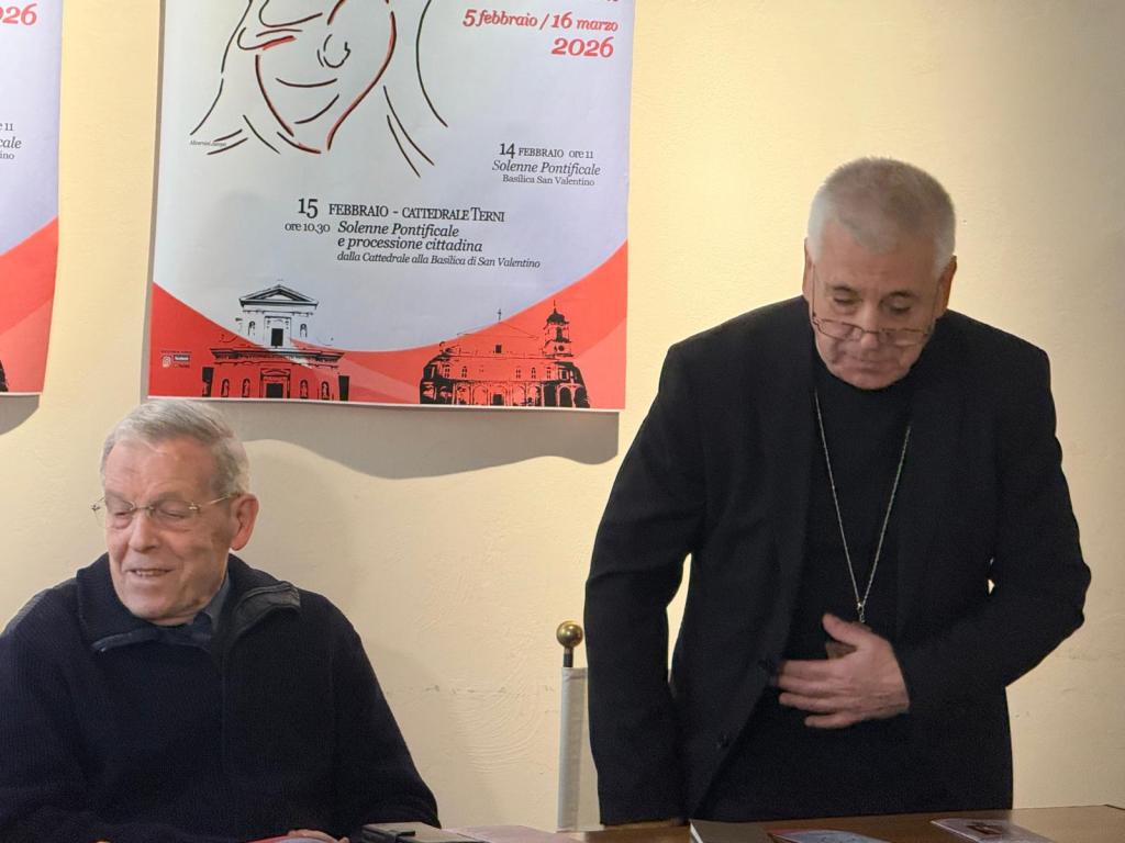 Two men at a table with event posters in the background, one smiling and the other adjusting his glasses.