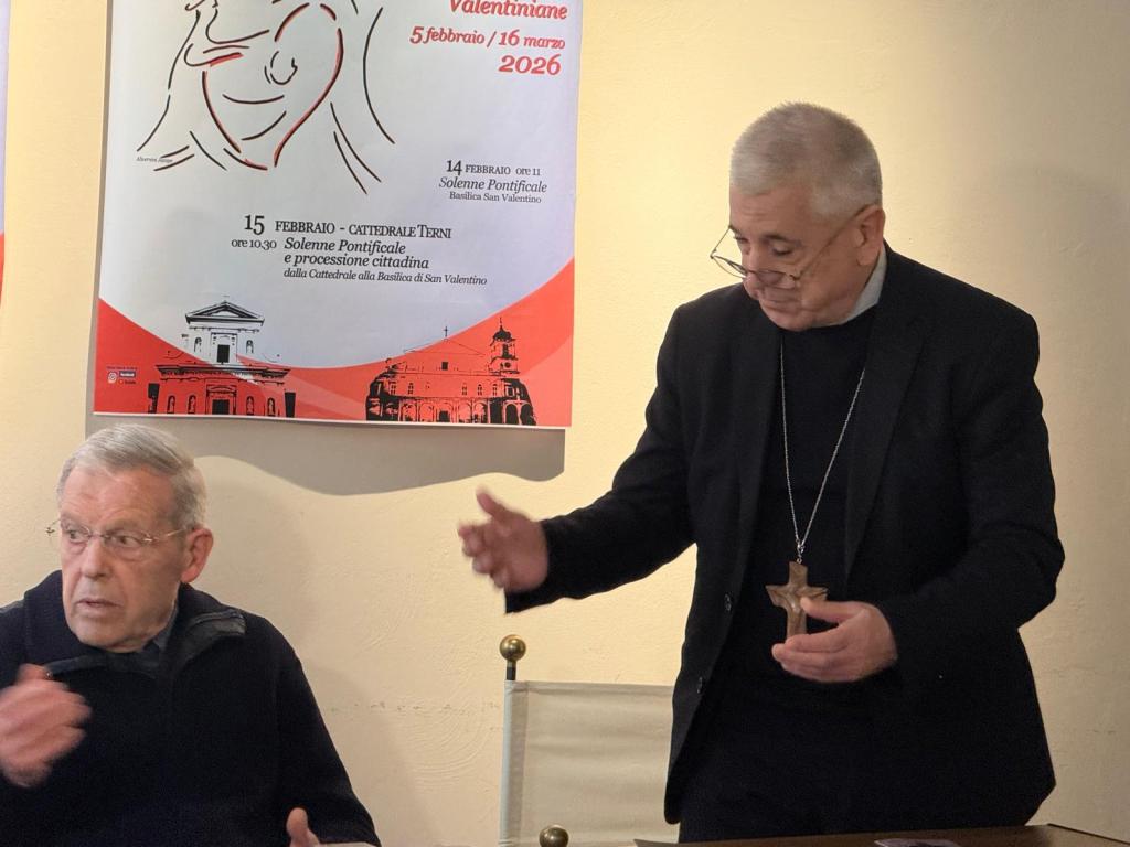 Two men are seated at a table in front of a poster advertising an event for Valentine's Day. One man, wearing glasses, appears to be listening attentively while the other man stands and gestures with a wooden cross in his hand.