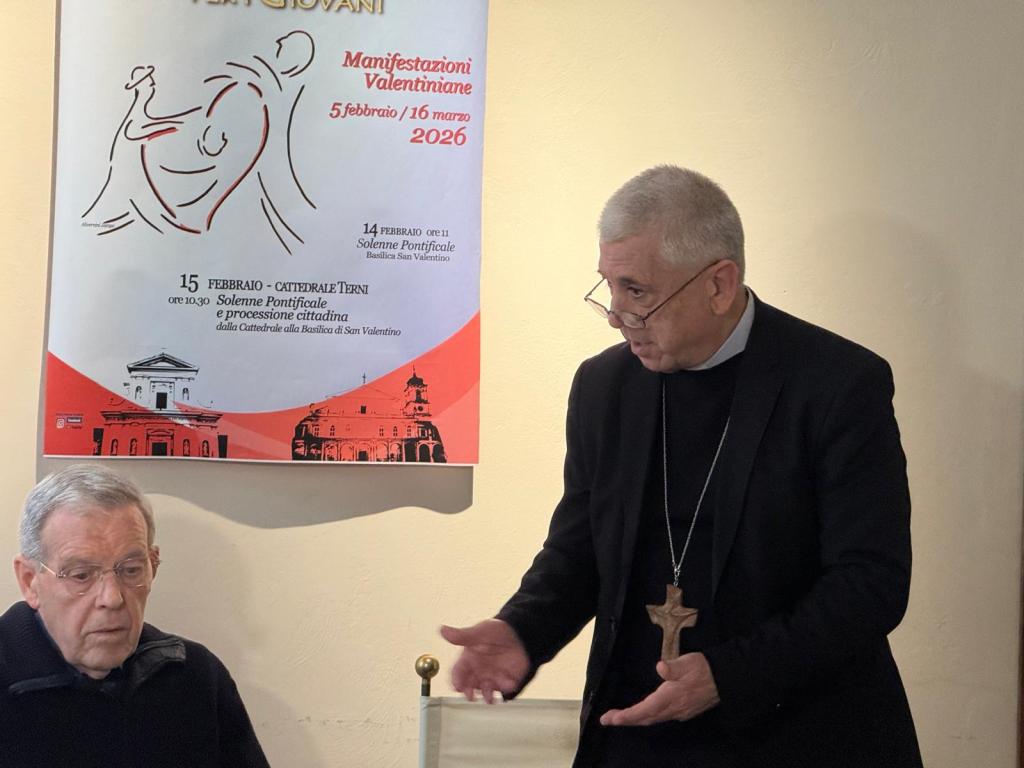 A man in clerical attire speaks to another seated man in a room, with a poster behind them detailing the Valentine's Day celebrations in Terni, Italy, scheduled for February and March 2026.