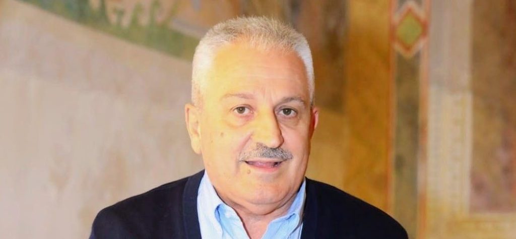 A middle-aged man with gray hair and a mustache, wearing a blue shirt and dark sweater, smiling at the camera with a blurred artistic background.