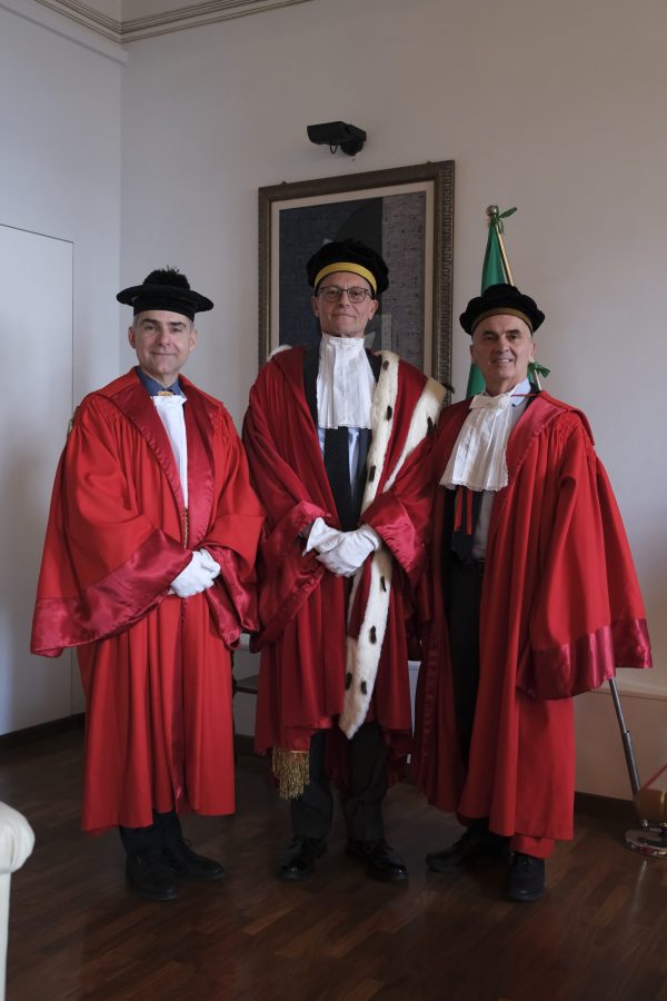 Three figures in ceremonial red robes and caps standing together in an indoor setting, with one wearing a fur-trimmed robe.