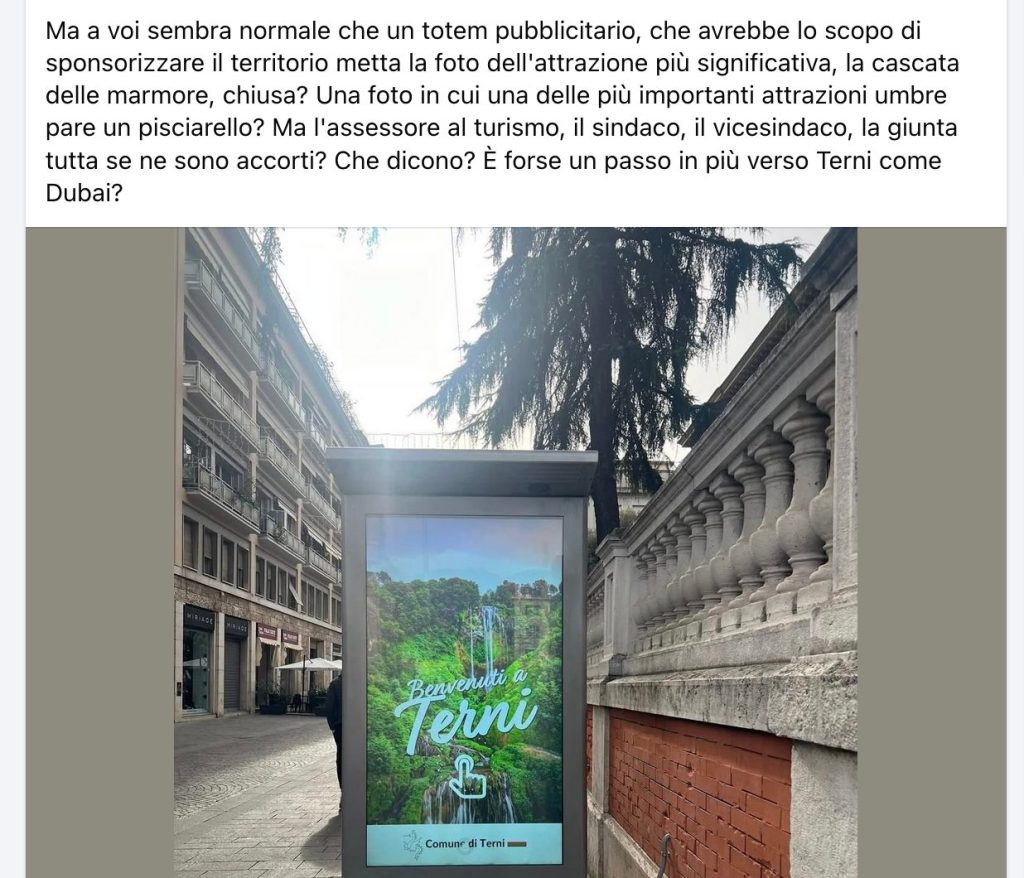 A large promotional totem displaying a welcome message for Terni, featuring a scenic image of a waterfall and lush greenery, set against a backdrop of urban buildings.